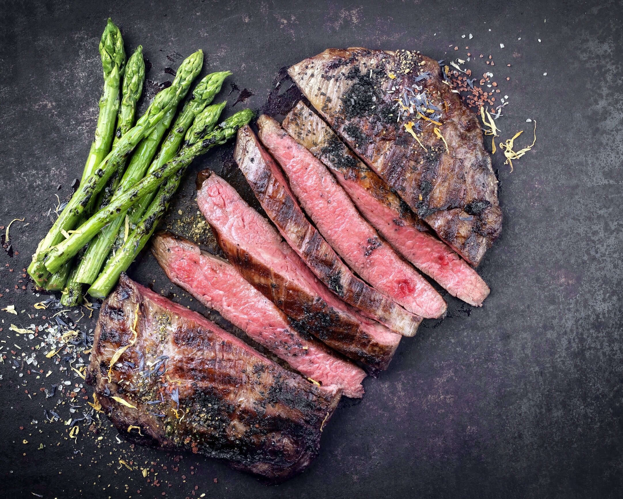 Grilled steak slices served alongside fresh asparagus on a dark textured surface, showcasing a delicious meal with vibrant colors and appealing presentation.