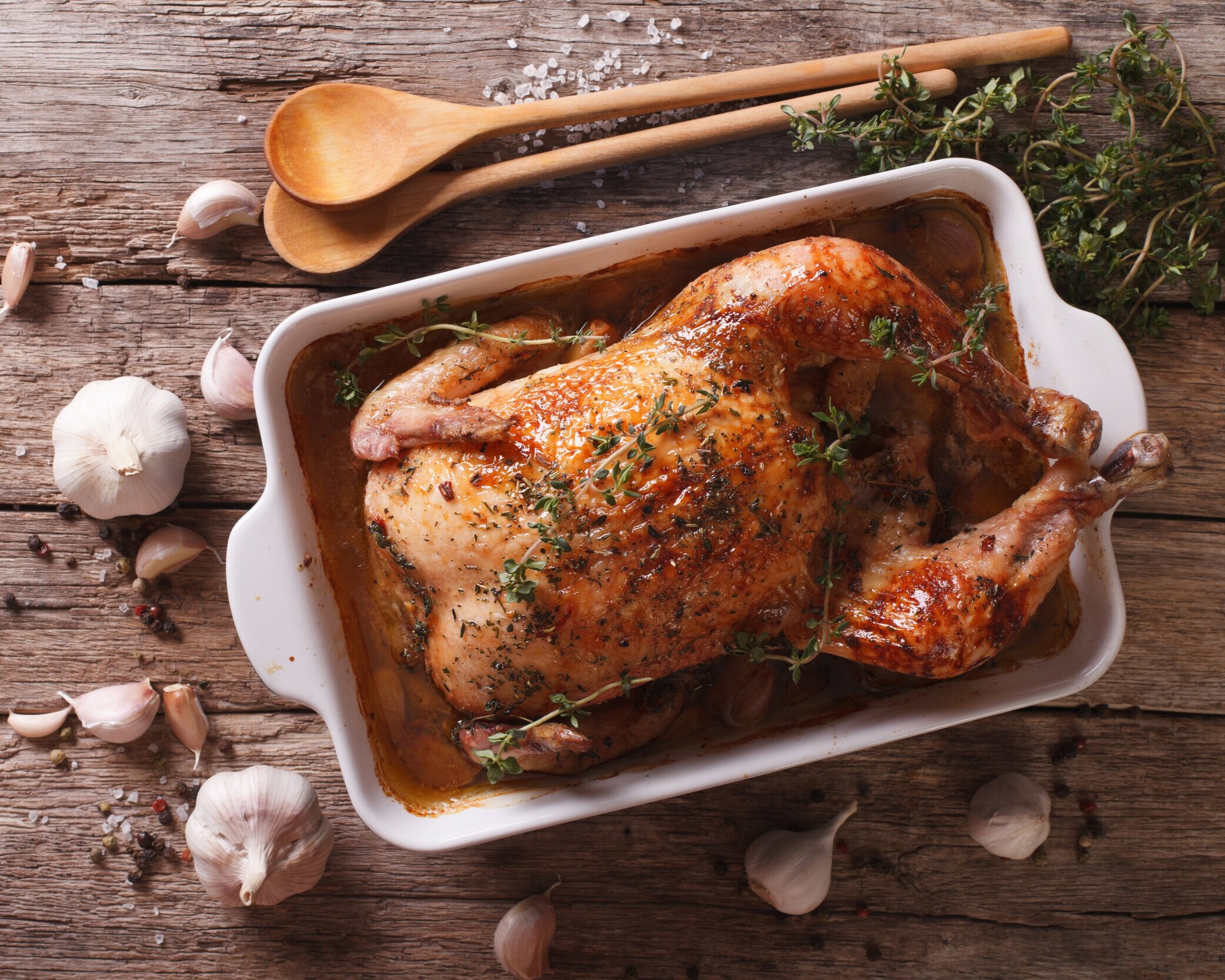 Roasted chicken garnished with fresh thyme, surrounded by garlic cloves and wooden utensils on a rustic wooden table. Perfect for a comforting home-cooked meal.
